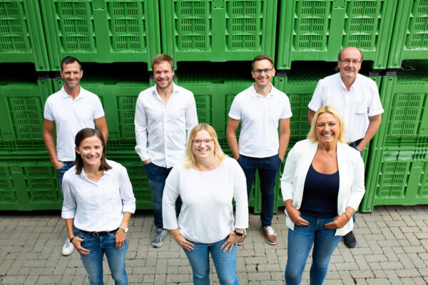 Das engagierte Europlast-Team posiert vor grünen Lagerhallen am Unternehmensstandort. Die Mitarbeiter:innen in weißer Berufskleidung und Freizeitkleidung zeigen Teamgeist und Professionalität in entspannter Atmosphäre.